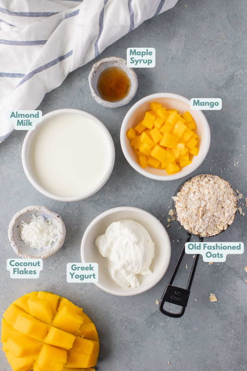 Ingredients for mango overnight oats in white bowls: oliold fashioned rolled oats, greek yogurt, almond milk, mango, maple syrup, unsweetened coconut flakes