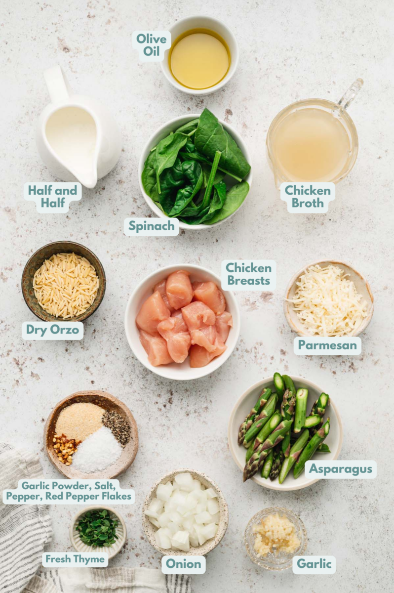 Ingredients for chicken orzo in small bowls on a marble background: chicken breasts, garlic powder, salt, pepper, red pepper flakes, olive oil, onion, garlic, asparagus, dry orzo, chicken broth, fresh thyme, half and half, parmesan, spinach