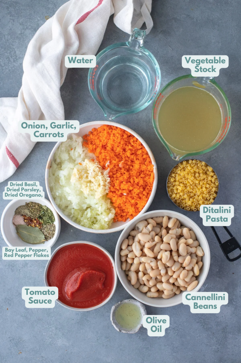 Ingredients for pasta fagioli in white bowls on a grey background: olive oil, cannellini beans, fresh herbs, garlic, onions, carrots, salt, pepper, red pepper flakes, short pasta, broth & water, tomato sauce
