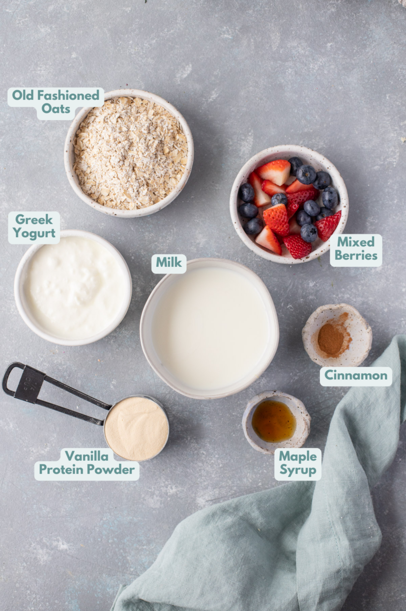 Ingredients for protein overnight oats in white bowls on a grey background: old fashioned rolled oats, nonfat greek yogurt, vanilla protein powder, almond milk, cinnamon, berries (strawberries and blueberries)