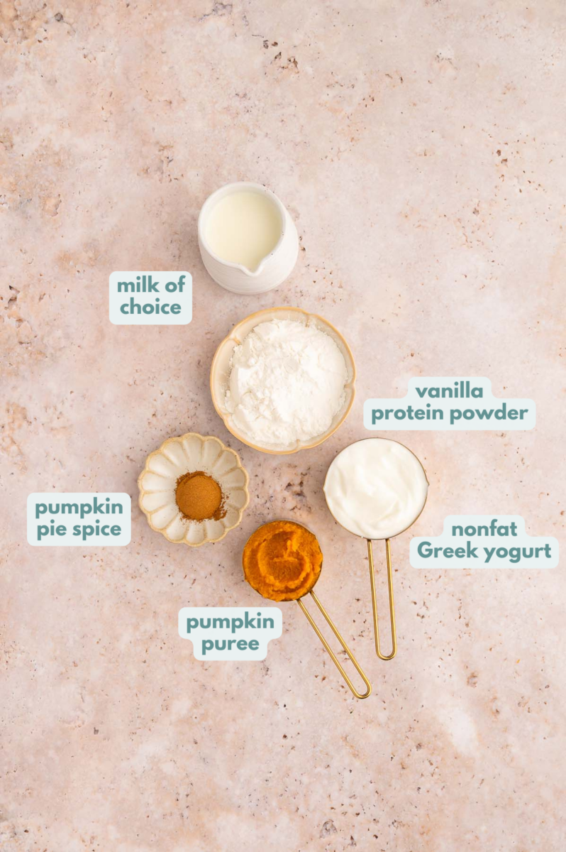 Ingredients for pumpkin protein smoothie in small bowls and measuring cups: milk of choice, vanilla protein powder, nonfat Greek yogurt, pumpkin puree, pumpkin pie spice.