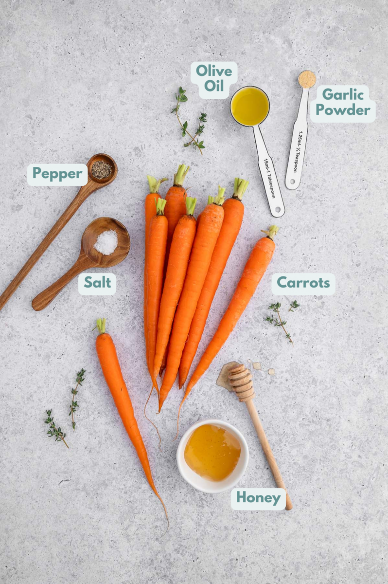 Ingredients for honey roasted carrots spread on a grey background: carrots, olive oil, salt, garlic powder, pepper, honey
