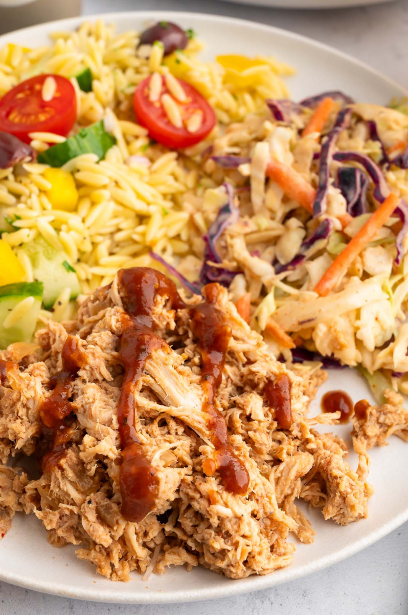 High Protein BBQ Chicken on a white plate with cole slaw and orzo salad in the background.