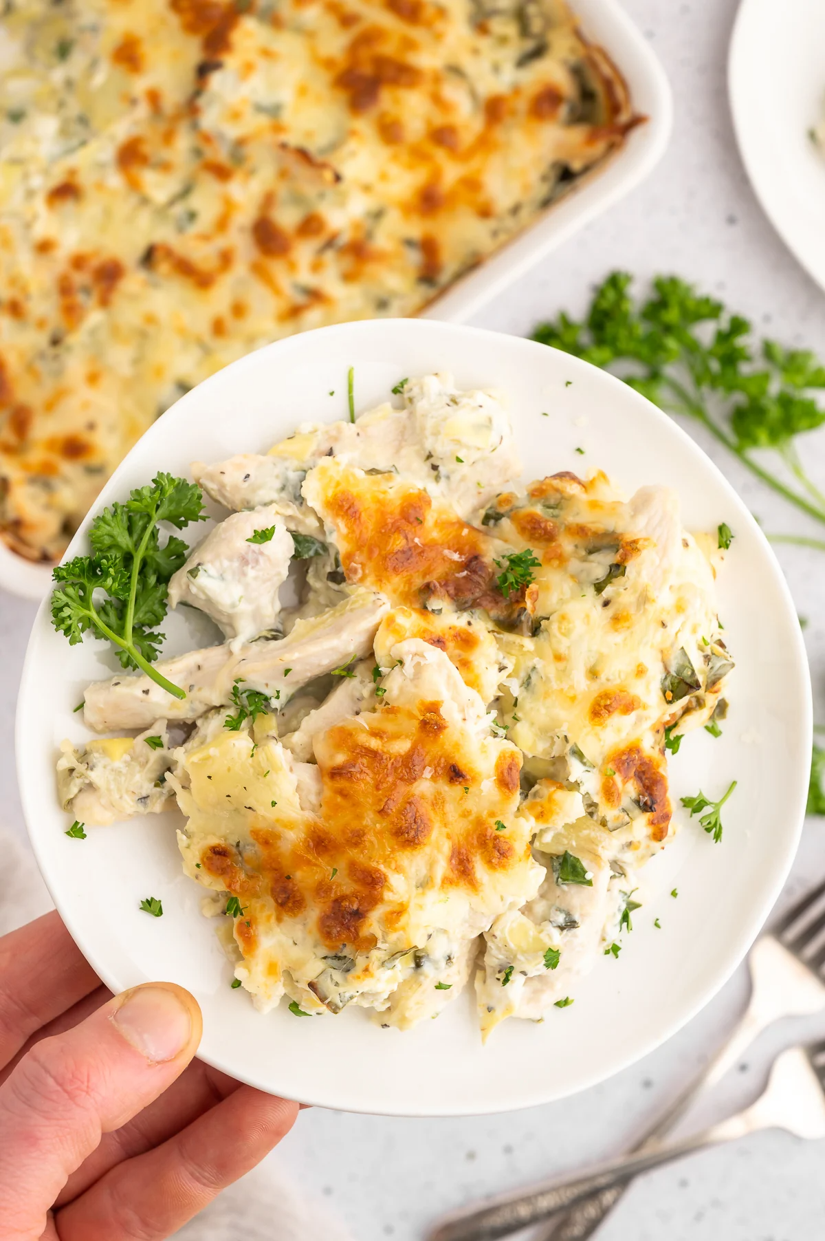 A hand holding up a plate with a serving of high protein spinach artichoke chicken and the baking dish of the casserole in the background.