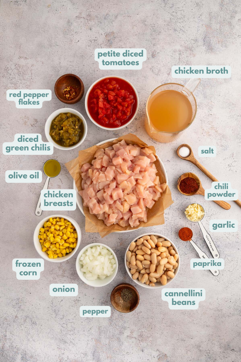 Chicken white bean chili ingredients in white bowls on a grey background: chicken breasts, cannellini beans, diced green chiles, frozen corn, chicken broth, onion, garlic, olive oil, chili powder, paprika, red pepper flakes, salt & pepper